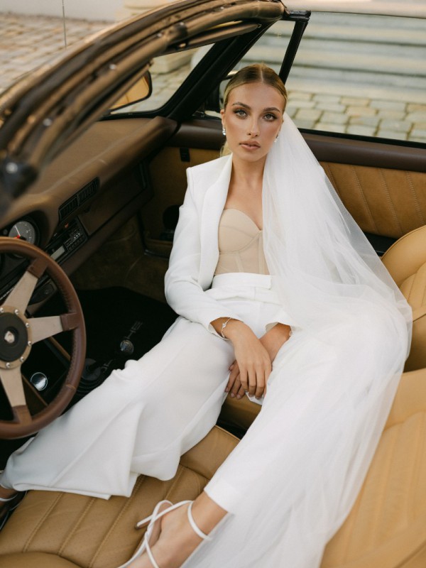 Braut im eleganten Hosenanzug mit Schleier sitzt stilvoll im Oldtimer am Tag ihrer Hochzeit