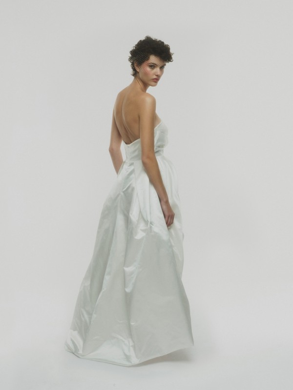 Modernes rückenfreies Brautkleid mit schulterfreiem Corsageteil und voluminösem Rock, Braut mit kurzem Haar und Fascinator blickt über die Schulter vor hellem Hintergrund