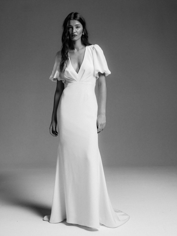 Schlichtes Clean-Brautkleid mit tiefem V-Ausschnitt und kurzen Puffärmeln, glatter weißer Stoff, Braut mit offenen Haaren im Schwarzweiß-Look vor neutralem Hintergrund