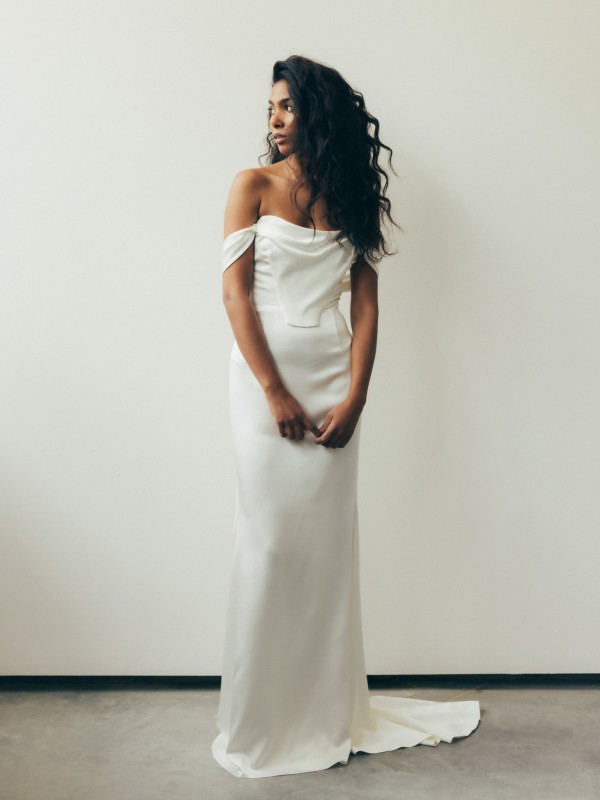 Clean-Brautkleid mit Off-Shoulder und moderner, glatter Silhouette, minimalistisch und elegant, Braut mit lockigem Haar vor weißer Wand